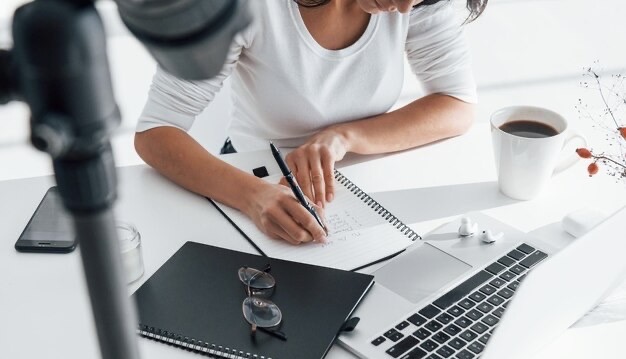 A girl writes something in a notepad, next to her is a laptop and a cup of tea