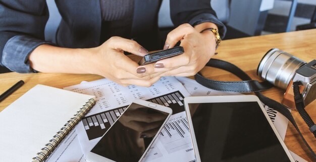 A woman holds a smartphone in her hands, papers and equipment are on the table next to her